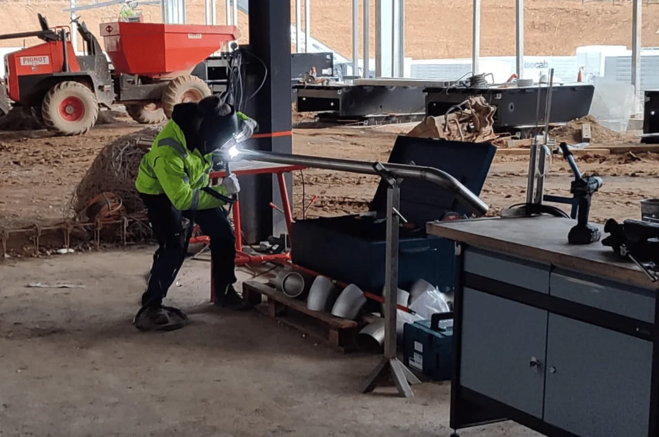 Na zdjęciu widać mężczyznę, który naprawia pewną rurę poprzez proces spawania. W specjalnym stroju ochronnym, wyposażony w maskę spawalniczą, rękawice i właściwe ubranie, koncentruje się na zadaniu.