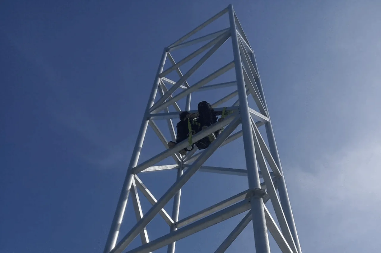 Na zdjęciu widać mężczyznę pracującego nad ziemią na metalowej konstrukcji. Jest ubrany w specjalny strój ochronny i wyposażony w niezbędne sprzęty do pracy na wysokościach, jak linia życia, pas bezpieczeństwa i kask.